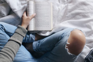 Woman in jeans sitting on a white bed and reading a book
