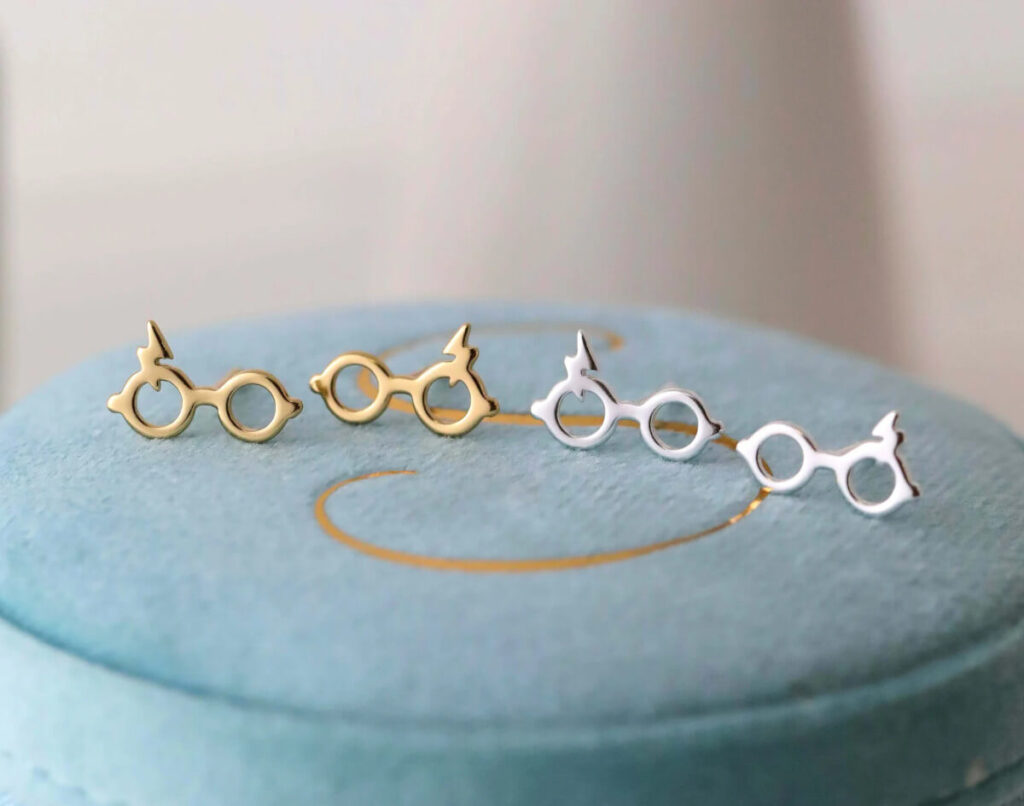 Two pairs of earrings shaped like glasses with a lightning bolt on top, one in silver and one in gold. By StudSparkleJewelry