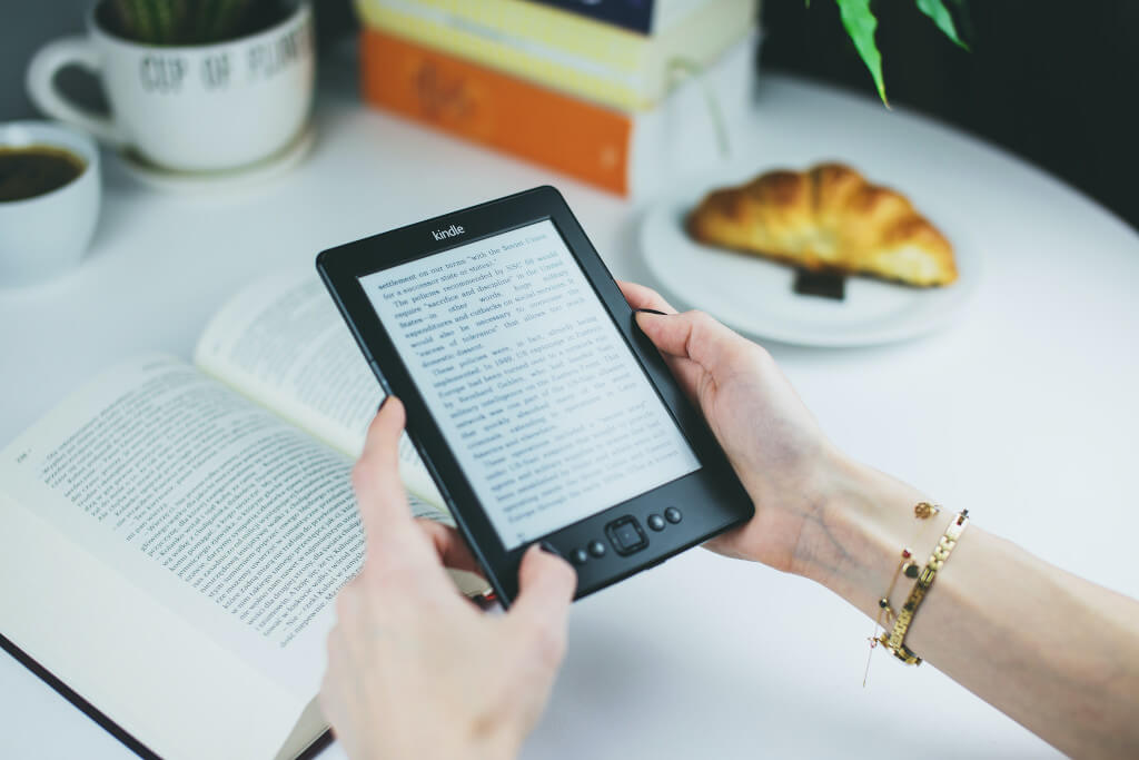 Hands holding a Kindle while seated at a table with books and a croissant