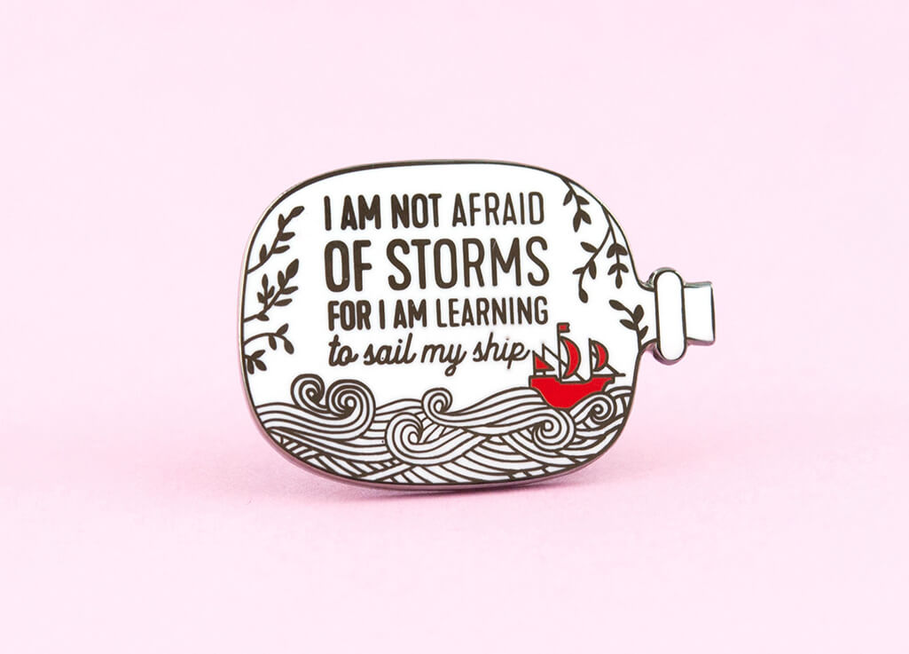Enamel pin shaped like a ship in the bottle, featuring a red ship on waves and the words "I am not afraid of storms for I am learning to sail my ship" by The Clever Clove
