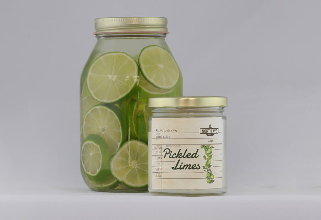 Glass candle reading "pickled limes", placed in front of a jar of limes and lime juice. Candle by North Ave Candles.
