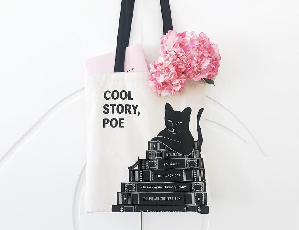 White tote with a black cat sitting on top of a stack of books, and text that reads "Cool Story, Poe." By Obvious State