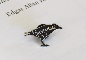 Black and silver enamel pin shaped like a raven, with the word "Nevermore" written across its body. By Literary Emporium