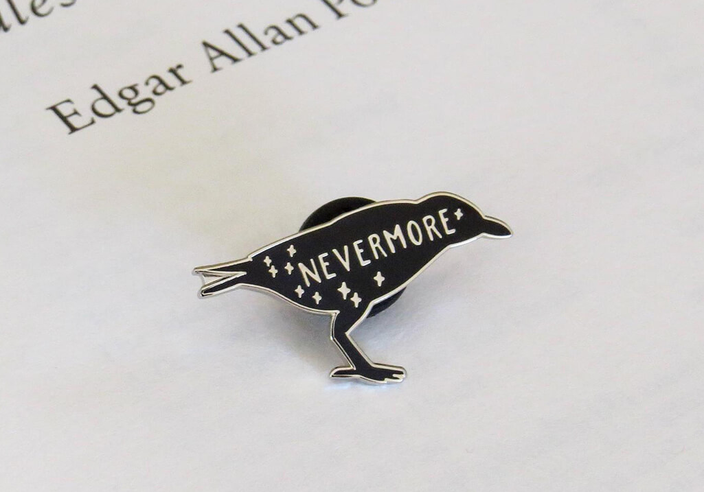 Black and silver enamel pin shaped like a raven, with the word "Nevermore" written across its body. By Literary Emporium
