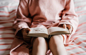 Girl wearing pink and sitting on a bed while reading a book