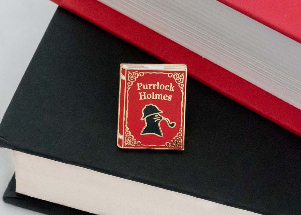 Red enamel pin shaped like a book with a silhouette of a cat smoking a pipe and the words "Purrlock Holmes"
