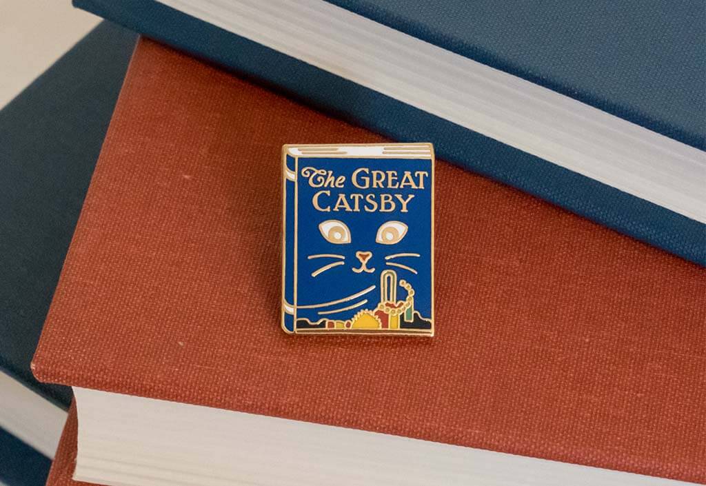 Blue enamel pin shaped like a book with a cat's eyes and whiskers. It reads "The Great Catsby"