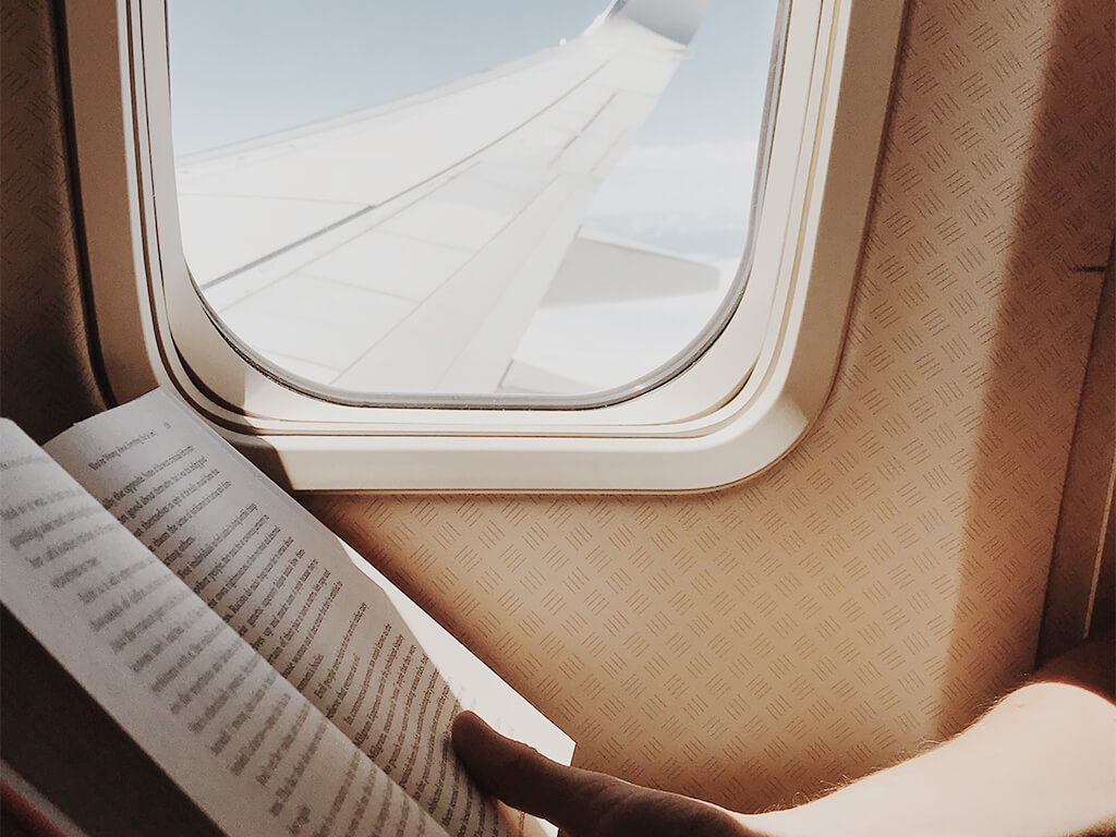 An open book next to an airplane window with a view of the plane's wing