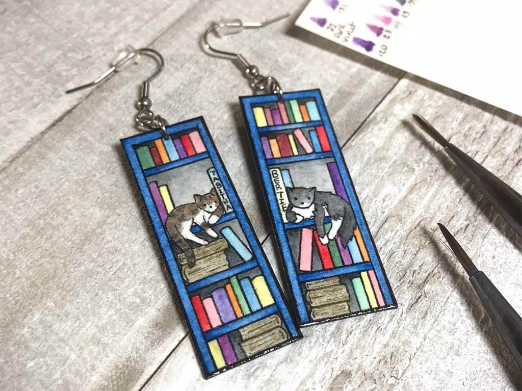 Earrings that look like a bookshelf with a cat sitting on one of the shelves by SeijaNeumyer