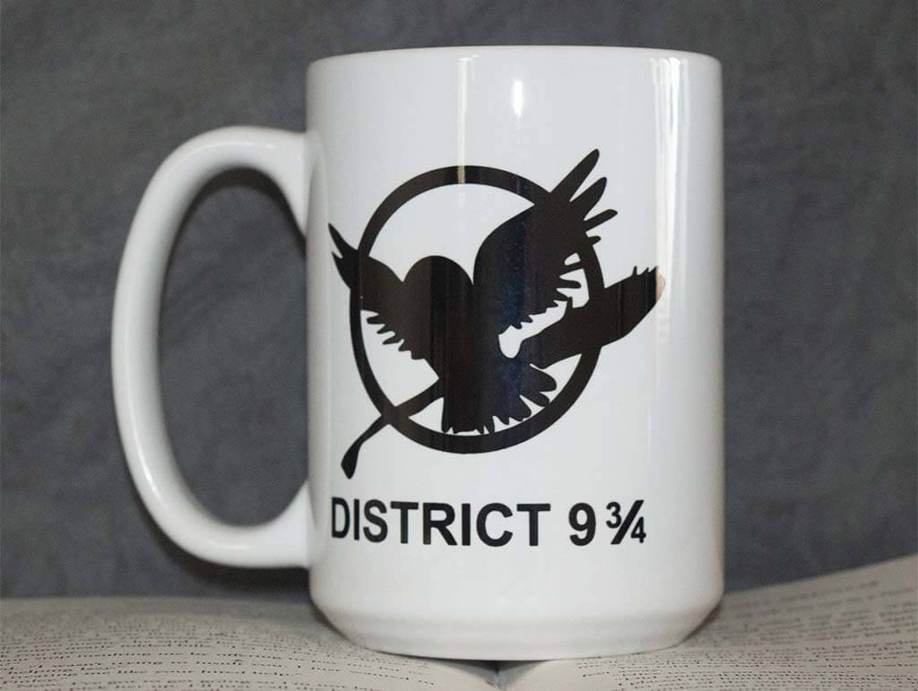 White mug with Hunger Games mockingjay symbol, expect replaced with an owl and broom and the words below read "District 9 3/4" by BookalorianDesigns