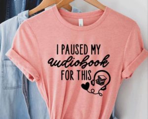 Pink tshirt with the words "I paused my audiobook for this" and a headphones and book icon. By MadeCuriouser
