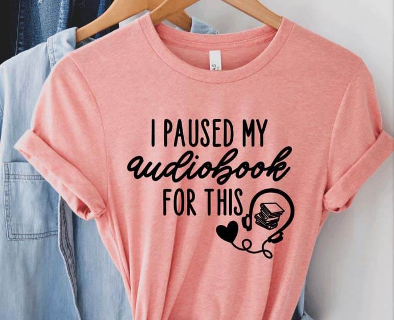 Pink tshirt with the words "I paused my audiobook for this" and a headphones and book icon. By MadeCuriouser