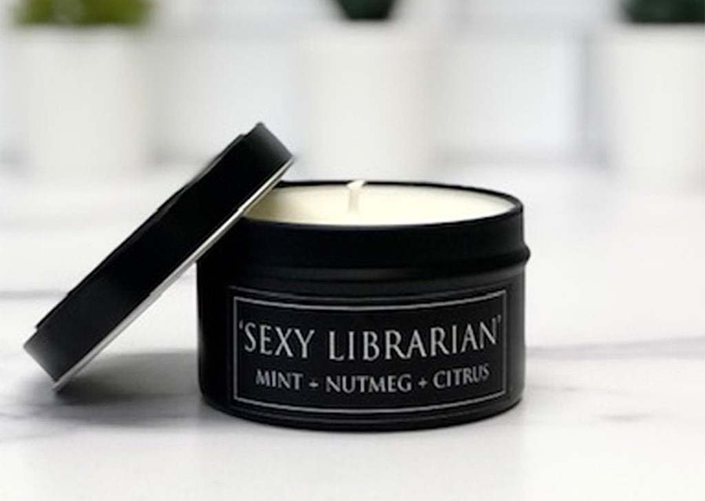 White candle in a black tin that reads "Sexy Librarian" by FlyPaperProducts