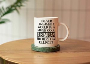 White mug with black text that reads "I never dreamed I would be a super cool librarian, but here I am, killing it" by Migoota