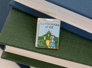 Book-shaped enamel pin with a cat in a Dorothy outfit. It reads "The Whiskers of Oz".
