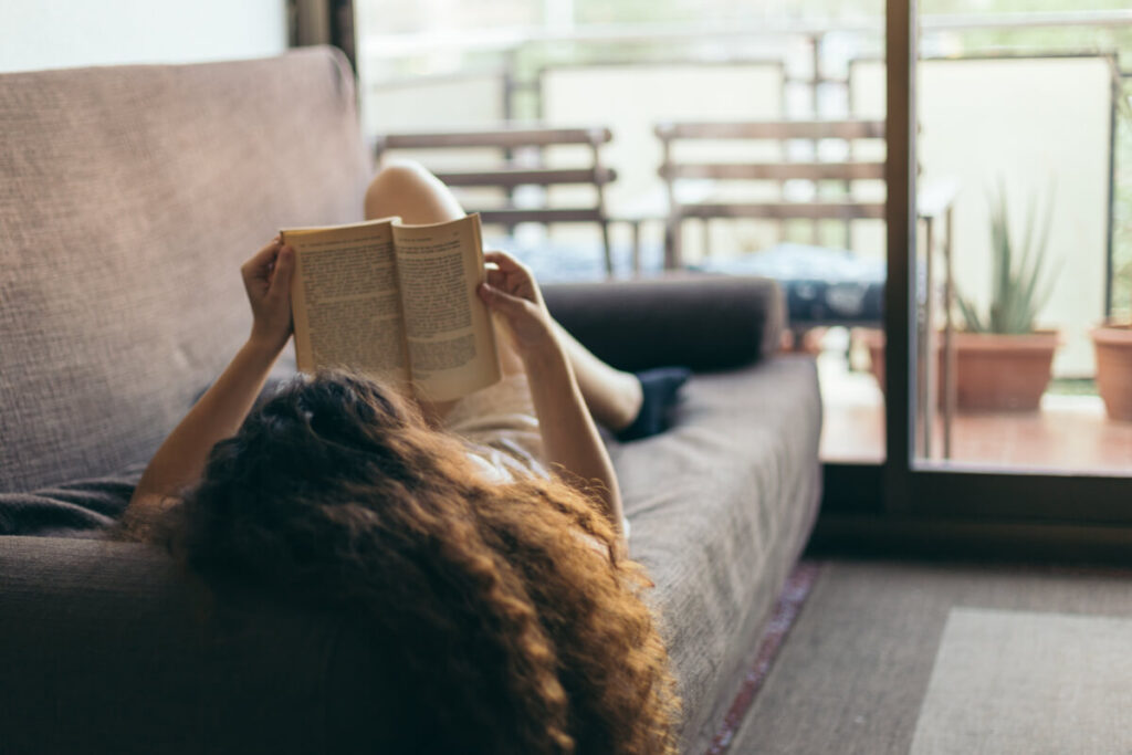 Girl with long curly hair lying down on a couch and reading an open book