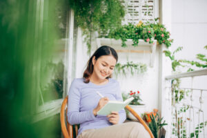 Woman surrounded by plants and sitting on a balcony, while writing in a journal
