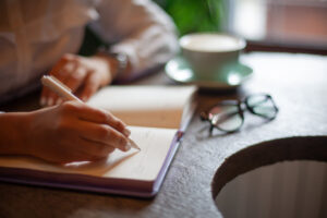 Woman writing in a journal while sitting in a coffee shop with eyeglasses and a coffee cup next to her