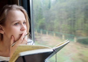 Woman looking thoughtful and writing in a journal next to a window on a train